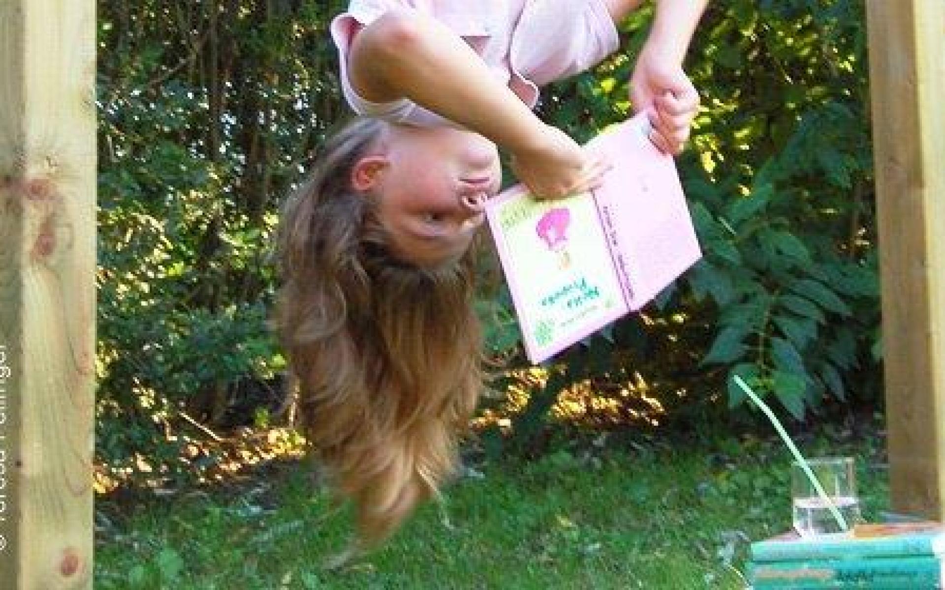 Mädchen hängt auf Stange mit dem Kopf nach unten und hat ein Buch in der Hand
